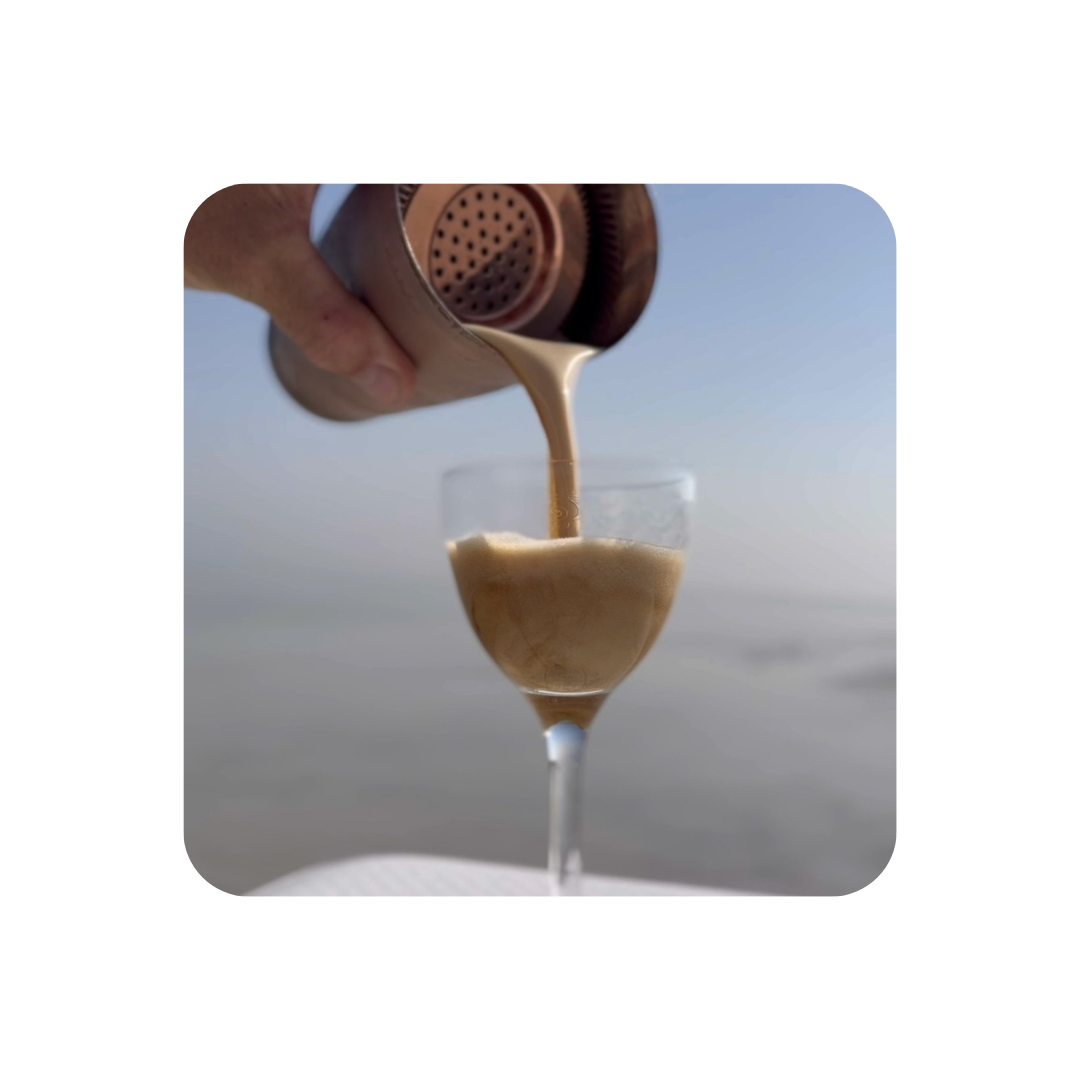 espresso martini being poured from cocktail shaker into glass with ocean in background
