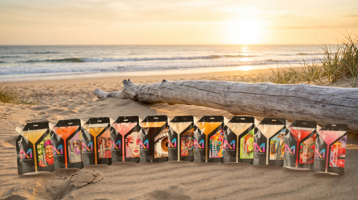All 11 MXTOLOGY cocktail pouches lined up on the sand at sunset beside driftwood