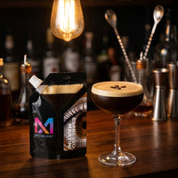 Espresso Martini MXTOLOGY cocktail pouch, shaken and activated with crema on top, beside a foamy coupe on a dark bar